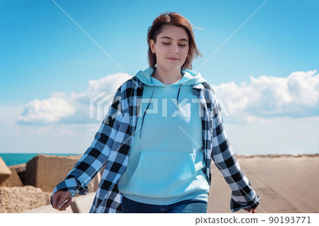 Teenager girl in light blue hoodie and plaid shirt walking outdoor, sunny day and blue sky 90193771