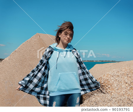 Teenager girl in light blue hoodie and plaid shirt against concrete blocks and blue sky 90193791