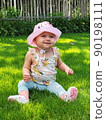 A small, happy, smiling baby in a pink hat, panama, sneakers sits on the grass in a village, a country house. Blue eyes. Against the backdrop of a fence 90198111