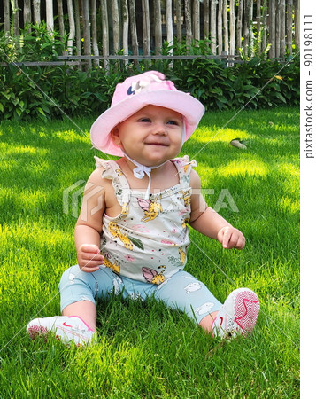 A small, happy, smiling baby in a pink hat, panama, sneakers sits on the grass in a village, a country house. Blue eyes. Against the backdrop of a fence A small, happy, smiling baby in a pink hat, panama, sneakers sits on the grass in a village, a country house. Blue eyes. Against the backdrop of a fence 90198111