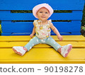 A small, smiling, happy, blue-eyed girl, a baby sits on a yellow-blue bench, the color of the Ukrainian flag. In the park. Pink hat, panama, pants, sneakers. Summer, spring, day 90198278