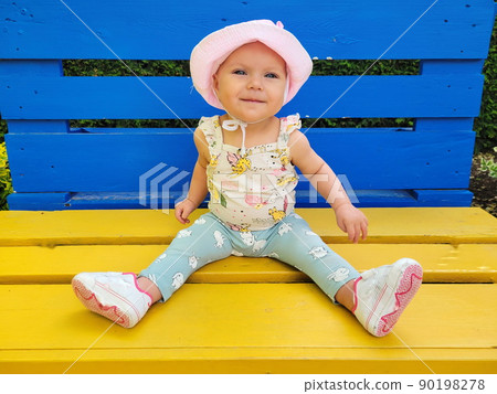 A small, smiling, happy, blue-eyed girl, a baby sits on a yellow-blue bench, the color of the Ukrainian flag. In the park. Pink hat, panama, pants, sneakers. Summer, spring, day 90198278