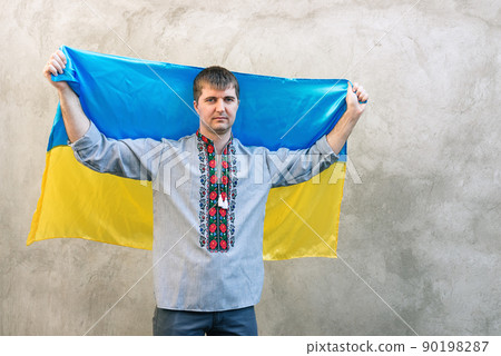 Front view of a young smiling man holding the blue and yellow national flag of Ukraine over his head. Man in traditional embroidered shirt on a background of textured concrete wall. Copy space Front view of a young smiling man holding the blue and yellow national flag of Ukraine over his head. Man in traditional embroidered shirt on a background of textured concrete wall. Copy space 90198287