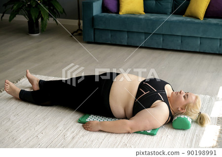 A fat obese woman is lying on the floor in a room on a massage needle massage mat and pillow. Relieving neck pain when the body is overweight. 90198991
