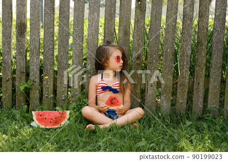 A curly-haired beautiful girl in red sunglasses is sitting on a lawn in the village and eating a watermelon, looking thoughtfully away A curly-haired beautiful girl in red sunglasses is sitting on a lawn in the village and eating a watermelon, looking thoughtfully away 90199023