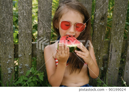 A curly-haired brunette girl bites a ripe red large piece of watermelon, closing her eyes from enjoying the sweetness of the taste. Refreshment on a hot summer day. A curly-haired brunette girl bites a ripe red large piece of watermelon, closing her eyes from enjoying the sweetness of the taste. Refreshment on a hot summer day. 90199024