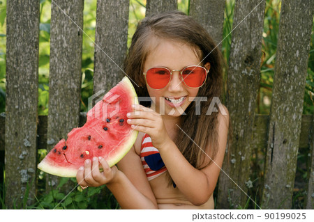 A laughing amusing girl holds a ripe red large piece of watermelon with bones in her hands. Enjoyment and refreshment on a hot summer day. A laughing amusing girl holds a ripe red large piece of watermelon with bones in her hands. Enjoyment and refreshment on a hot summer day. 90199025