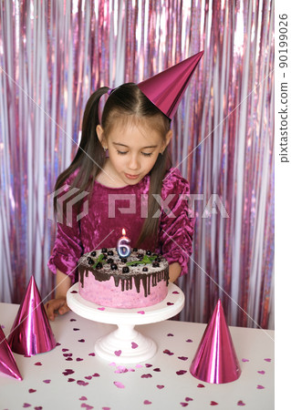 A cute little girl in a pink dress with a festive hat on her head blows out a candle in the shape of the number 6 on the cake. The child celebrates his birthday. A cute little girl in a pink dress with a festive hat on her head blows out a candle in the shape of the number 6 on the cake. The child celebrates his birthday. 90199026