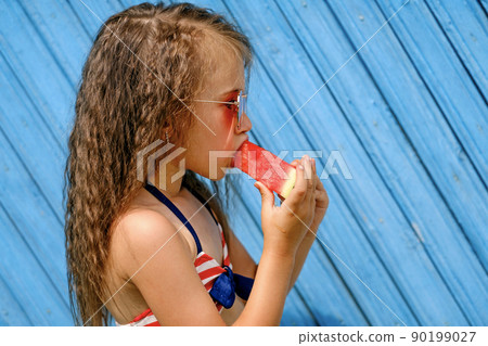 Profile of a beautiful curly-haired brunette girl biting a slice of juicy red watermelon. A child in sunglasses on a blue background of wooden planks Profile of a beautiful curly-haired brunette girl biting a slice of juicy red watermelon. A child in sunglasses on a blue background of wooden planks 90199027