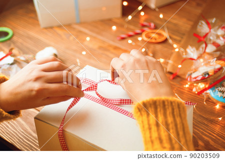 Woman's hands in knitted sweater tie red ribbon bow on white gift box. New Year's mood. Garland lights, gingerbread and decorations in background. Surprise for Birthday or Christmas. 90203509