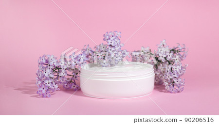 White jar of moisturizing face cream with pink cap isolated on pink background with lilac flowers. eco cosmetology or skin care concept. White jar of moisturizing face cream with pink cap isolated on pink background with lilac flowers. eco cosmetology or skin care concept. 90206516