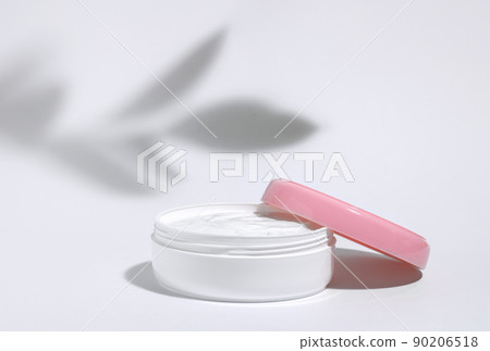 The background is cosmetic. White jar of moisturizing face cream with pink cap isolated on white background with shadow from plant branch. Cosmetology or skin care concept. Copy space The background is cosmetic. White jar of moisturizing face cream with pink cap isolated on white background with shadow from plant branch. Cosmetology or skin care concept. Copy space 90206518