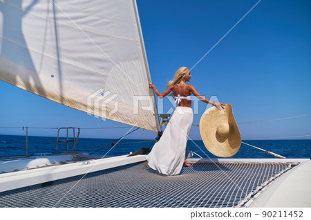 Beautiful young blond woman in bikini standing on catamaran at sunny summer day 90211452