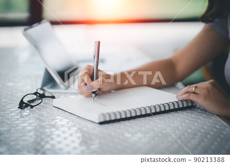 closeup hands of business woman working on laptop and using pen making notices in her notebook on wooden table with a cup of coffee. Business concept 90213388