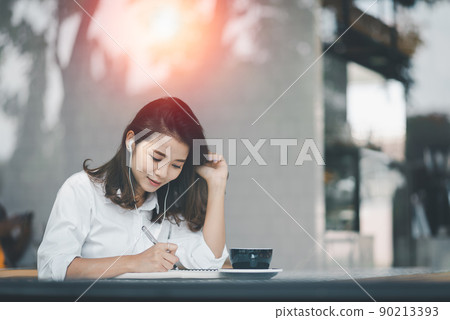 Asian woman sitting and using a pen to write on a paper notebook with a laptop computer and wearing earphones. Work from anywhere concept. Work from home concept. Asian woman sitting and using a pen to write on a paper notebook with a laptop computer and wearing earphones. Work from anywhere concept. Work from home concept. 90213393