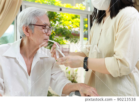 Asian caregiver take care of senior woman give drinking water from a glass,feeding water using a straw to prevent choking at nursing home,thirsty elderly patient sucking water through straw for safety 90213538