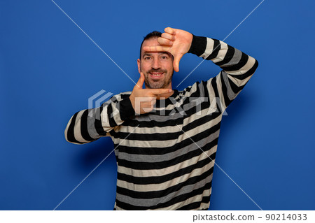 Handsome bearded happy male blink with eye and looking through a frame formed by his hands. Attractive man make frame by fingers, isolated on blue background with copy space for text. People concept. Handsome bearded happy male blink with eye and looking through a frame formed by his hands. Attractive man make frame by fingers, isolated on blue background with copy space for text. People concept. 90214033