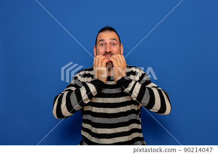Latino man with his hands on his mouth feeling authentic terror, he bites his nails to try to calm his fears. Isolated on blue studio background. Latino man with his hands on his mouth feeling authentic terror, he bites his nails to try to calm his fears. Isolated on blue studio background. 90214047