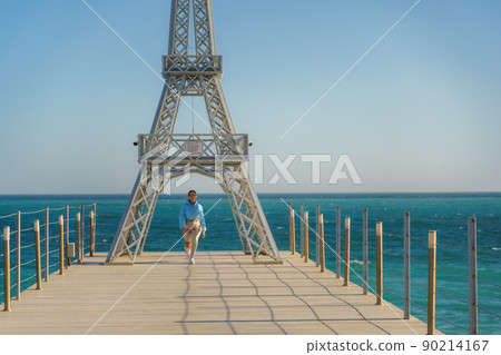 Large model of the Eiffel Tower on the beach. A woman walks along the pier towards the tower, wearing a blue jacket and white jeans. 90214167