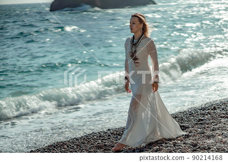 Middle aged woman looks good with blond hair, boho style in white long dress on the beach decorations on her neck and arms. 90214168