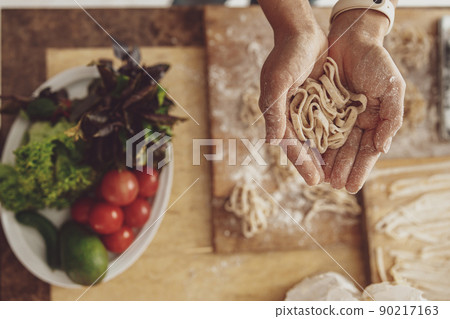 Homemade noodles in women's hands over a kitchen board with flour and vegetables on a plate 90217163