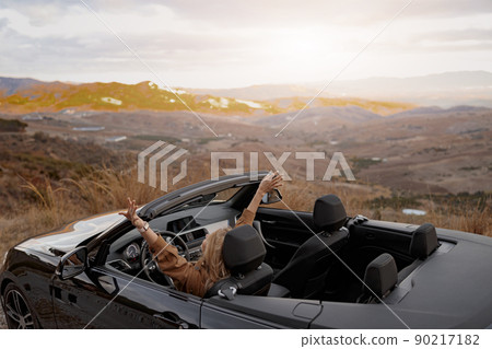 Rear of young pretty happy female holding hand up feeling free sitting in convertible car 90217182