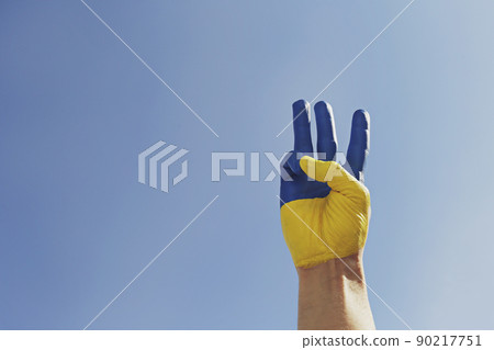 Man's hand lit by sunshine painted in colors of Ukrainian national flag showing a traditional trident hand gesture as a symbol of support and solidarity with Ukraine. Copy space, close up, background. Man's hand lit by sunshine painted in colors of Ukrainian national flag showing a traditional trident hand gesture as a symbol of support and solidarity with Ukraine. Copy space, close up, background. 90217751
