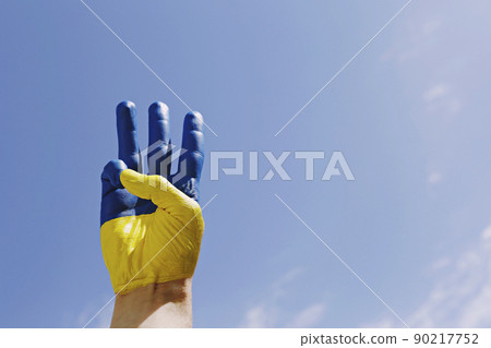 Man's hand lit by sunshine painted in colors of Ukrainian national flag showing a traditional trident hand gesture as a symbol of support and solidarity with Ukraine. Copy space, close up, background. Man's hand lit by sunshine painted in colors of Ukrainian national flag showing a traditional trident hand gesture as a symbol of support and solidarity with Ukraine. Copy space, close up, background. 90217752