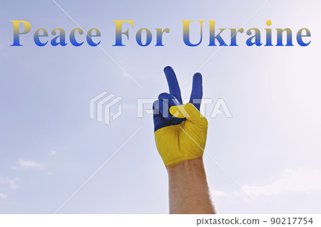 Peace for Ukraine slogan written over blue sky background. Man's hand painted in colors of Ukrainian national flag showing a peace gesture as sign of support and solidarity with Ukraine. Copy space. Peace for Ukraine slogan written over blue sky background. Man's hand painted in colors of Ukrainian national flag showing a peace gesture as sign of support and solidarity with Ukraine. Copy space. 90217754