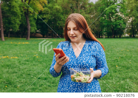 Portrait of attractive caucasian smiling woman eating salad. Happy young person using mobile phone to give positive rate of customer service mobile app. Healthy food and technology banner 90218076