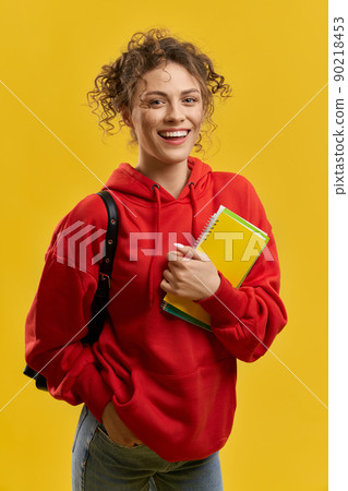 Cute girl with backpack, looking at camera, while holding notes indoor. Front view of female student standing with hand in pocket, smiling, isolated on yellow studio background. Concept of studying.  90218453