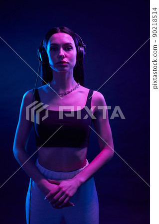 Confident woman in sporty outfit standing with folded arms in studio. Portrait view of serious girl wearing black headphones looking at camera, illuminated with magenta light. Concept of lifestyle. Confident woman in sporty outfit standing with folded arms in studio. Portrait view of serious girl wearing black headphones looking at camera, illuminated with magenta light. Concept of lifestyle. 90218514