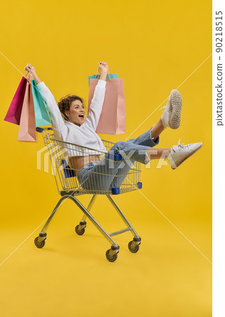Cheerful girl holding colorful bags, while riding in shopping cart indoor. Side view of happy lady having fun with trolley, while shopping, isolated on yellow studio background. Concept of happiness. Cheerful girl holding colorful bags, while riding in shopping cart indoor. Side view of happy lady having fun with trolley, while shopping, isolated on yellow studio background. Concept of happiness. 90218515