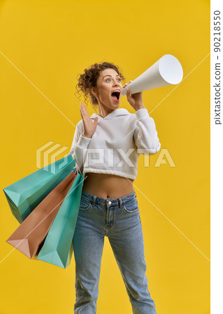 Caucasian woman shouting into megaphone, while shopping inside. Female shopper holding white bullhorn, loudly screaming, isolated on orange studio background, copy space. Concept of advertisement. 90218550
