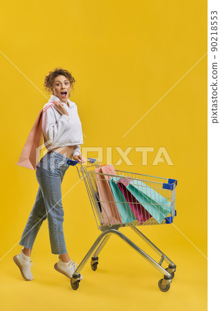 Shocked woman carrying shopping cart, with bright paper bags inside. Side view of surprised girl with shopping trolley, standing on tiptoe, isolated on orange studio background. Concept of shopping. Shocked woman carrying shopping cart, with bright paper bags inside. Side view of surprised girl with shopping trolley, standing on tiptoe, isolated on orange studio background. Concept of shopping. 90218553