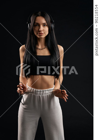 Young woman in headphones looking at the camera in studio. Portrait of brunette in black tank top standing straight, with earphones wire wrapped around hands, isolated on black. Concept of music fan. Young woman in headphones looking at the camera in studio. Portrait of brunette in black tank top standing straight, with earphones wire wrapped around hands, isolated on black. Concept of music fan. 90218554