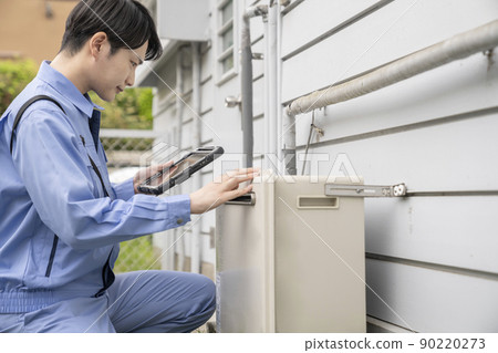 A man in work clothes inspecting a gas water heater installed outdoors in a house 90220273