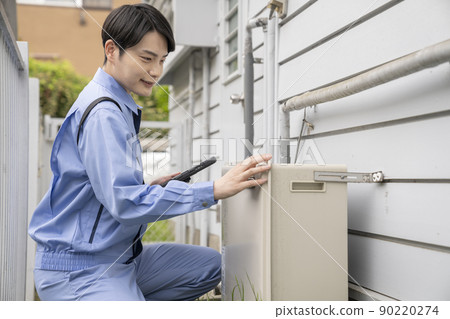 A man in work clothes inspecting a gas water heater installed outdoors in a single-family house 90220274