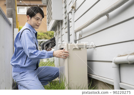 A man in work clothes inspecting a gas water heater installed outdoors in a house 90220275