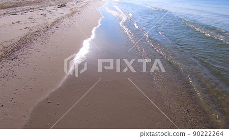 Waves of clean transparent water roll on the sandy beach of the sea shore. Person walking along the sea sandy beach. POV. Sun glare in the water. Seascape, waterscape background. Waves of clean transparent water roll on the sandy beach of the sea shore. Person walking along the sea sandy beach. POV. Sun glare in the water. Seascape, waterscape background. 90222264