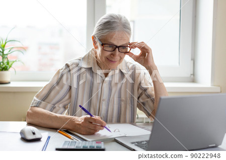 Intelligent caucasian senior woman adjust her glasses and look at laptops screen with a pen in hand, indoor, selective focus. 90222948