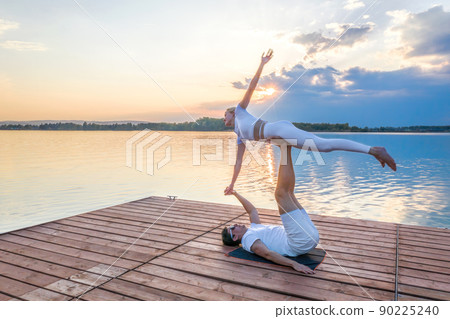 Beautiful young couple doing acro yoga on the pier against sunset. Man lying on the pier and balancing woman in his feet. 90225240