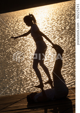 Beautiful young couple doing acro yoga on the pier against sunset. Man lying on the pier and balancing woman in his feet. Beautiful young couple doing acro yoga on the pier against sunset. Man lying on the pier and balancing woman in his feet. 90225241
