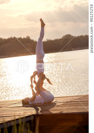 Beautiful young couple doing acro yoga on the pier against sunset. Man lying on the pier and balancing woman in his feet. Beautiful young couple doing acro yoga on the pier against sunset. Man lying on the pier and balancing woman in his feet. 90225258
