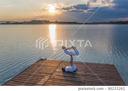 Beautiful young couple doing acro yoga on the pier against sunset. Man lying on the pier and balancing woman in his feet. Beautiful young couple doing acro yoga on the pier against sunset. Man lying on the pier and balancing woman in his feet. 90225261