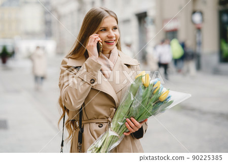 Beautiful girl in a brown coat. Woman in a spring city. Lady with bouquet of flowers 90225385