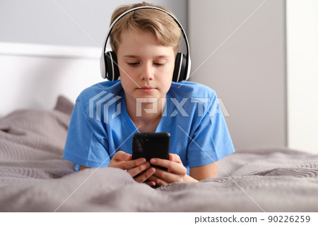 Close-up portrait of a cute boy in headphones. A teenager with a smartphone lies on a bed at home 90226259