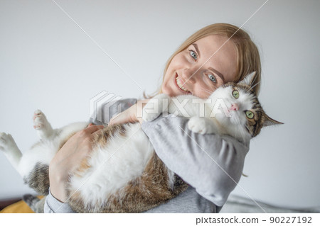 Portrait of young blonde happy 30s woman holding cute tabby cat with green eyes. Female hugging cat in hands 90227192