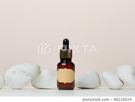 A brown glass bottle with a paper label and a pipette on a white table in the middle of the stones. 90228024