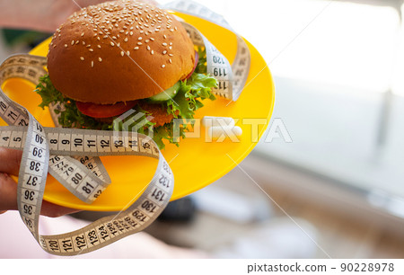 Woman holding unhealthy burger with measuring tape in kitchen. 90228978